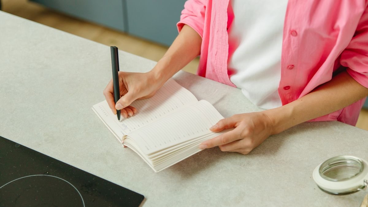 hand writing a grocery list in a notebook on a kitchen counter